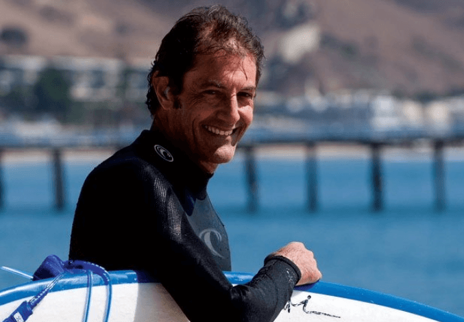 Shaun Tomson, world champion South African surfer, smiling beside his surfboard at the beach.
