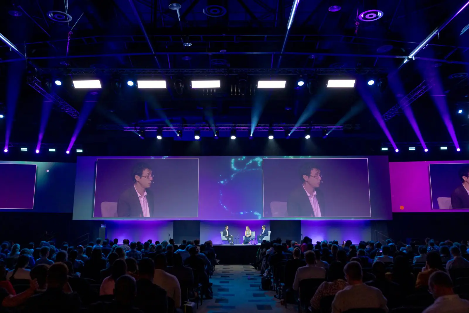 A large audience watching a fireside chat under purple stage lighting